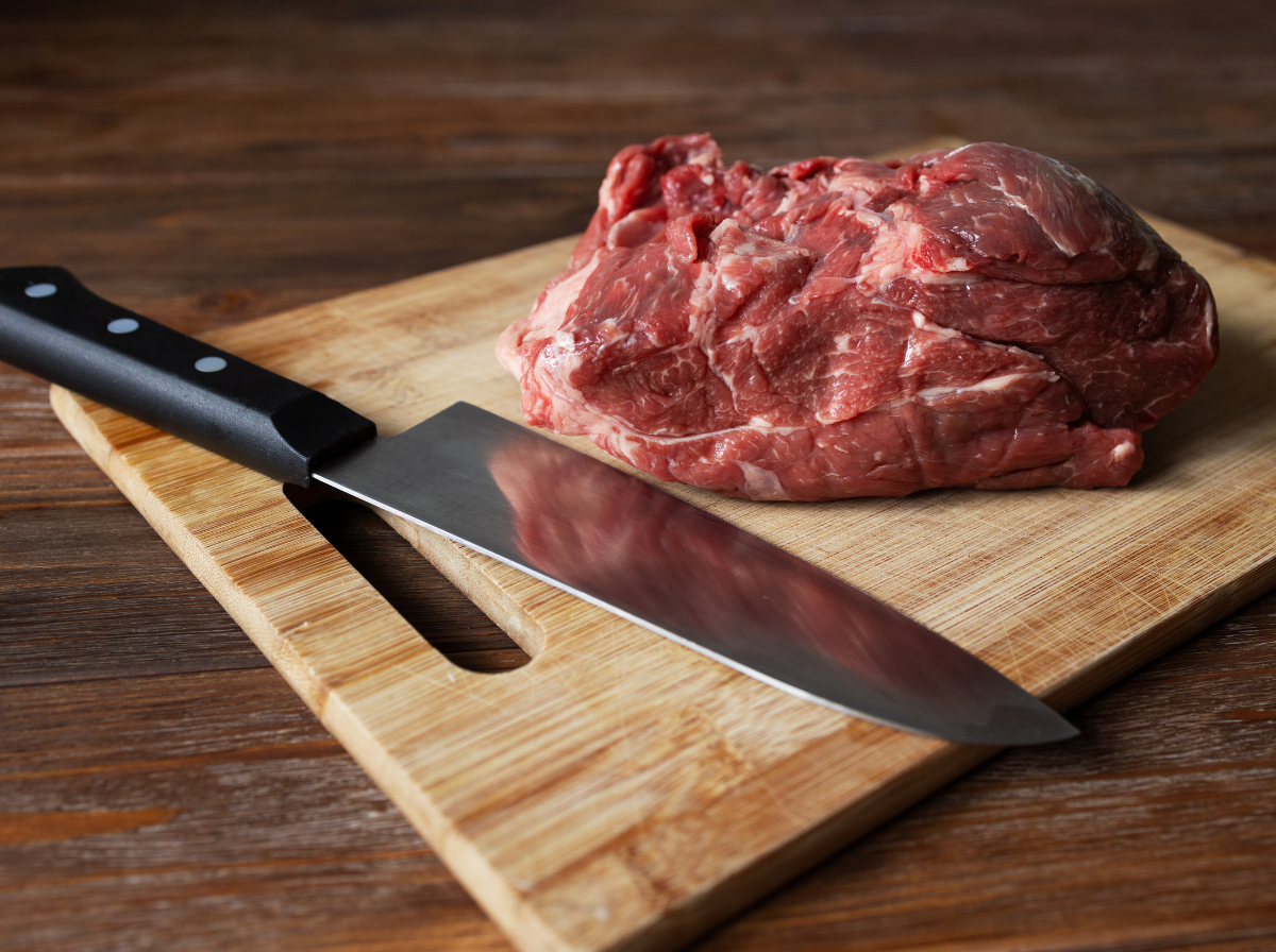 A sharp stainless steel chef's knife resting beside a raw beef roast on a wooden cutting board.